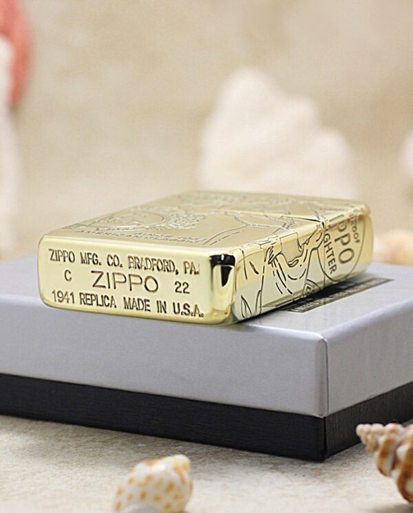KZ161-05 Gold Zippo lighter with engraved design resting on a gray gift box, seashells scattered around, markings read 'ZIPPO MFG. CO. BRADFORD, PA' and '1941 REPLICA MADE IN USA'