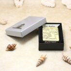 Zippo lighter with a vintage 1941 Replica design in an open black presentation box on a sandy surface with several seashells nearby.