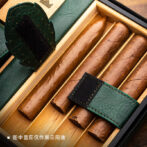 Cigars arranged in a wooden humidor compartment with green leather accents and a black strap, ready for storage.
