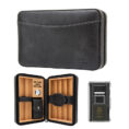 Black leather cigar travel case, opened to show wooden cigar dividers, a small humidification gauge, and a strap-secured pouch inside; lighter sits nearby.