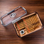 Open glass-trosted humidor lid hinged to the side, showing neatly arranged cigars inside next to a small digital hygrometer on a wooden surface.