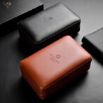Two leather zip cases, black and brown, with subtle embossed logos on a dark textured surface.