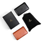 Set of leather wallets in black and tan with a black drawstring pouch and a black box on a white background, showing brand embossing on each item.