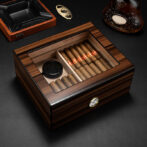 Wooden cigar humidor with a glass top open to reveal cigars arranged in two compartments and a small round humidifier plate; a hygrometer is mounted on the front, with an ashtray and bottle visible on a dark surface nearby.