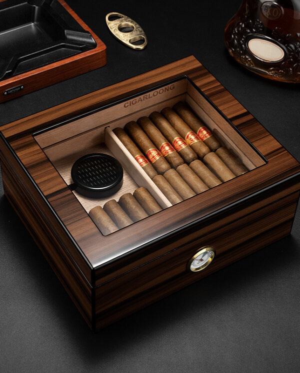 CH003-01 Wooden cigar humidor with a glass top open to reveal cigars arranged in two compartments and a small round humidifier plate; a hygrometer is mounted on the front, with an ashtray and bottle visible on a dark surface nearby.