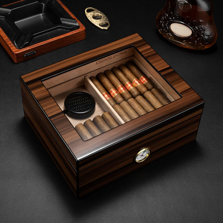 Wooden cigar humidor with a glass top open to reveal cigars arranged in two compartments and a small round humidifier plate; a hygrometer is mounted on the front, with an ashtray and bottle visible on a dark surface nearby.