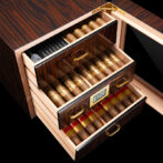Wooden cigar humidor with two open drawers packed with cigars; a digital hygrometer display visible on the front, and a small humidifier tray in the top drawer.