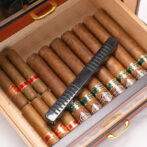 Open wooden humidor with many cigars and a black rectangular humidifier resting across them.