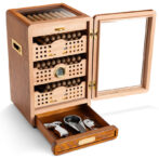 Open wooden cigar humidor with glass lid; displays cigars on shelves and a drawer containing cigar cutters and a lighter.
