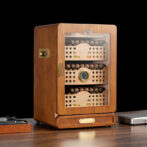 Wooden cigar humidor with glass front revealing multiple cigar shelves and a hygrometer on the middle shelf surface.
