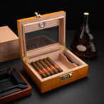 Open wooden cigar humidor with six cigars neatly arranged inside, a lock on the front, and a bottle of cognac nearby.