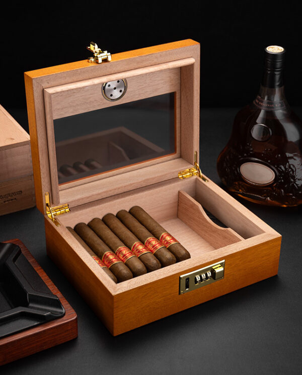 Open wooden cigar humidor with six cigars neatly arranged inside, a lock on the front, and a bottle of cognac nearby.