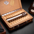 Opened wooden cigar box with multiple divider slots; a brown-and-white striped strap rests across the tray; a bottle sits nearby on the right.