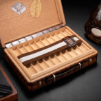 Opened wooden cigar box with multiple divider slots; a brown-and-white striped strap rests across the tray; a bottle sits nearby on the right.