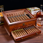 Open wooden cigar humidor with cigars arranged in rows; drawer pulled out showing more cigars.