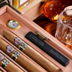 Cigars in a wooden humidor with a brass hygrometer and a black humidifier; whiskey bottle and glass in the background.