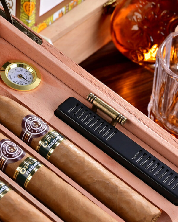 Cigars in a wooden humidor with a brass hygrometer and a black humidifier; whiskey bottle and glass in the background.