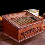 Polished wooden cigar humidor opened to reveal a row of cigars resting in a built-in tray with a key lock and decorative carving on the front panel.
