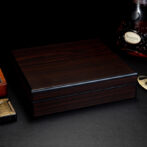 Polished dark wood jewelry or cigar box on a black textured surface with a cognac bottle nearby and a gold lighter in the corner.