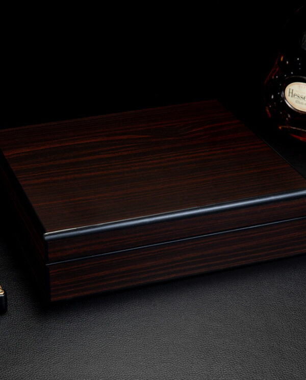 CH018-02 Polished dark wood jewelry or cigar box on a black textured surface with a cognac bottle nearby and a gold lighter in the corner.
