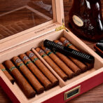 Cigar humidor open with multiple cigars lined up, a cutter resting on top, and a bottle of cognac nearby.