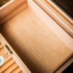 Interior of a light wood cigar humidor drawer with brass hardware and a small round hygrometer in the corner, empty inside.