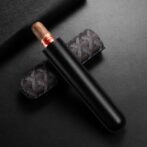 Brown cigar with red band in a black leather cigar sleeve laid on a dark background; a patterned case sits beneath the cigar.