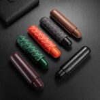 Six leather eyeglass cases in black, brown, burgundy, green, red, and gray with patterned fabric exteriors on a dark surface.