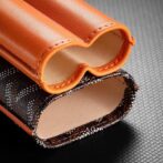 Close-up of two orange leather sleeves with stitched edges, resting on a textured black surface. A patterned fabric panel is visible beneath the sleeves.