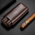 Brown leather cigar case beside a partially smoked cigar on a dark textured surface; a premium smoking accessory set up for display.