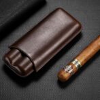Brown leather cigar case beside a partially smoked cigar on a dark textured surface; a premium smoking accessory set up for display.