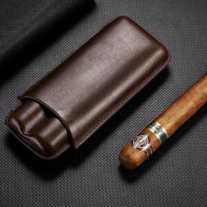 Brown leather cigar case beside a partially smoked cigar on a dark textured surface; a premium smoking accessory set up for display.