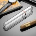 Luxurious smoking accessories on a glossy surface: a clear crystal cigar tube, a brown cigar, a wristwatch, and a CigarLong magazine in the background.