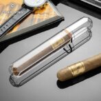 Crystal cigar tube on a reflective surface with a brown cigar nearby and a magazine and watch in the background.