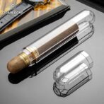 Transparent crystal cigar tube with a brown cigar inside, placed on a reflective black surface; brand logo visible in the corner.