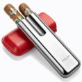 Stainless steel double-blade cigar cutter with two cigars resting on top and a red leather case behind it, angled diagonally.