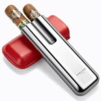 Stainless steel double-blade cigar cutter with two cigars resting on top and a red leather case behind it, angled diagonally.