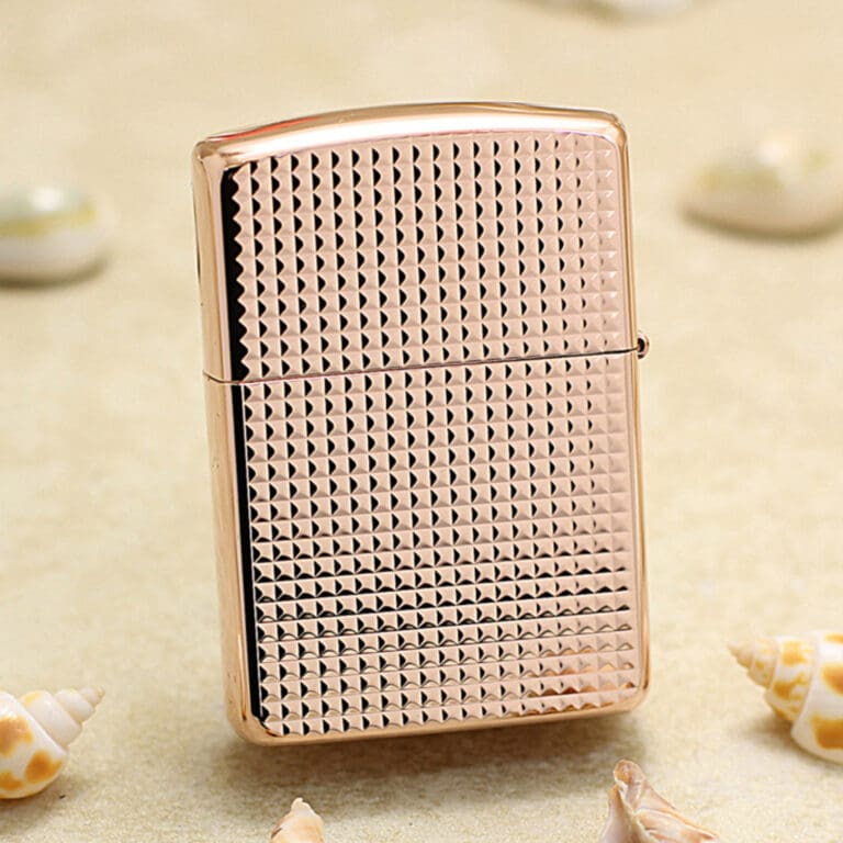 CurveS_01 Rose-gold Zippo-style lighter with a textured geometric pattern on a sandy surface, surrounded by seashells nearby.