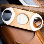 Wooden double-hole cigar cutter with metal rims resting on a brown leather case, a cigar lying behind it.