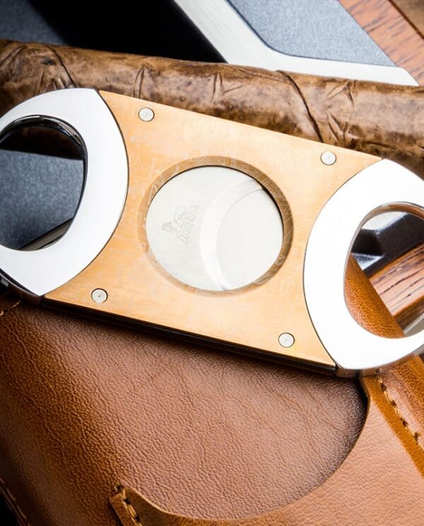HY-3305L-02 Wooden double-hole cigar cutter with metal rims resting on a brown leather case, a cigar lying behind it.