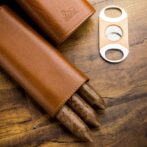 Leather cigar case with three brown cigars partially exposed, beside a metal double-blade cigar cutter on a wooden surface.