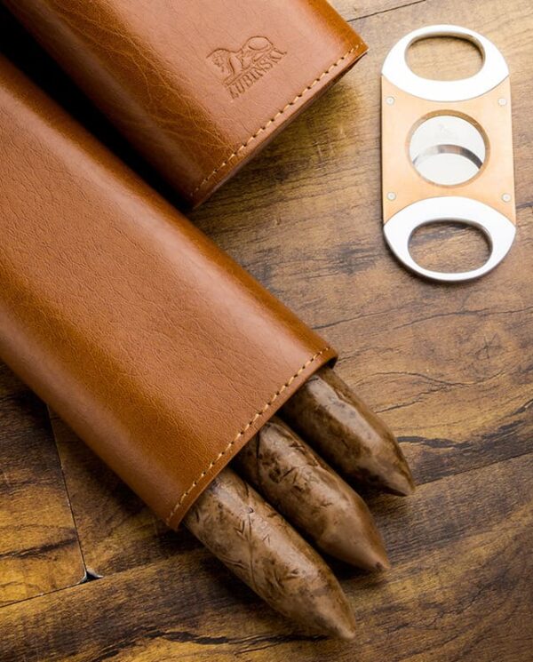 HY-3305L-03 Leather cigar case with three brown cigars partially exposed, beside a metal double-blade cigar cutter on a wooden surface.