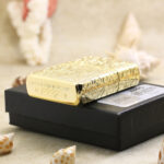 Golden arabesque lighter resting on its box with base and decorated side in view