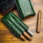 Green crocodile-pattern leather cigar cases with two protruding cigars and a metal cutter on a wooden surface.