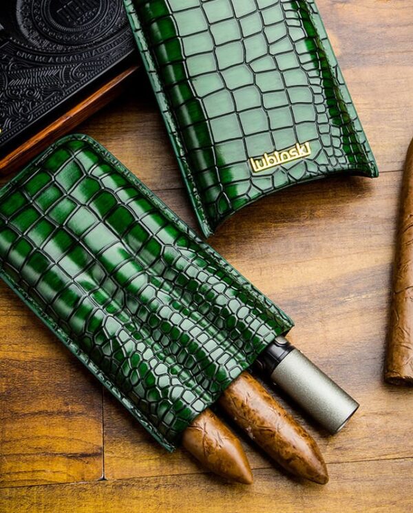 Green crocodile-pattern leather cigar cases with two protruding cigars and a metal cutter on a wooden surface.