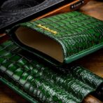 Open green crocodile-embossed leather clutch with tan interior and gold brand accent on a wooden surface.