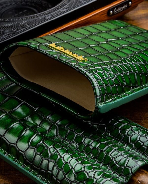 Open green crocodile-embossed leather clutch with tan interior and gold brand accent on a wooden surface.