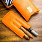 Two brown cigars partially exposed in orange woven leather cigar sleeves on a wooden desk surface.