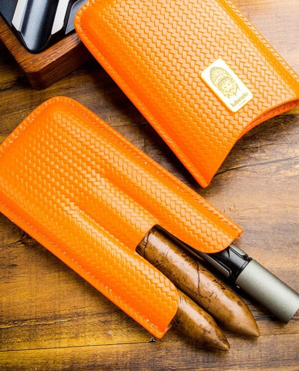 Two brown cigars partially exposed in orange woven leather cigar sleeves on a wooden desk surface.