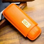 Orange woven leather case with a gold badge on the front, resting on a wood surface. It appears to be a compact accessory or sheath with stitched edges.